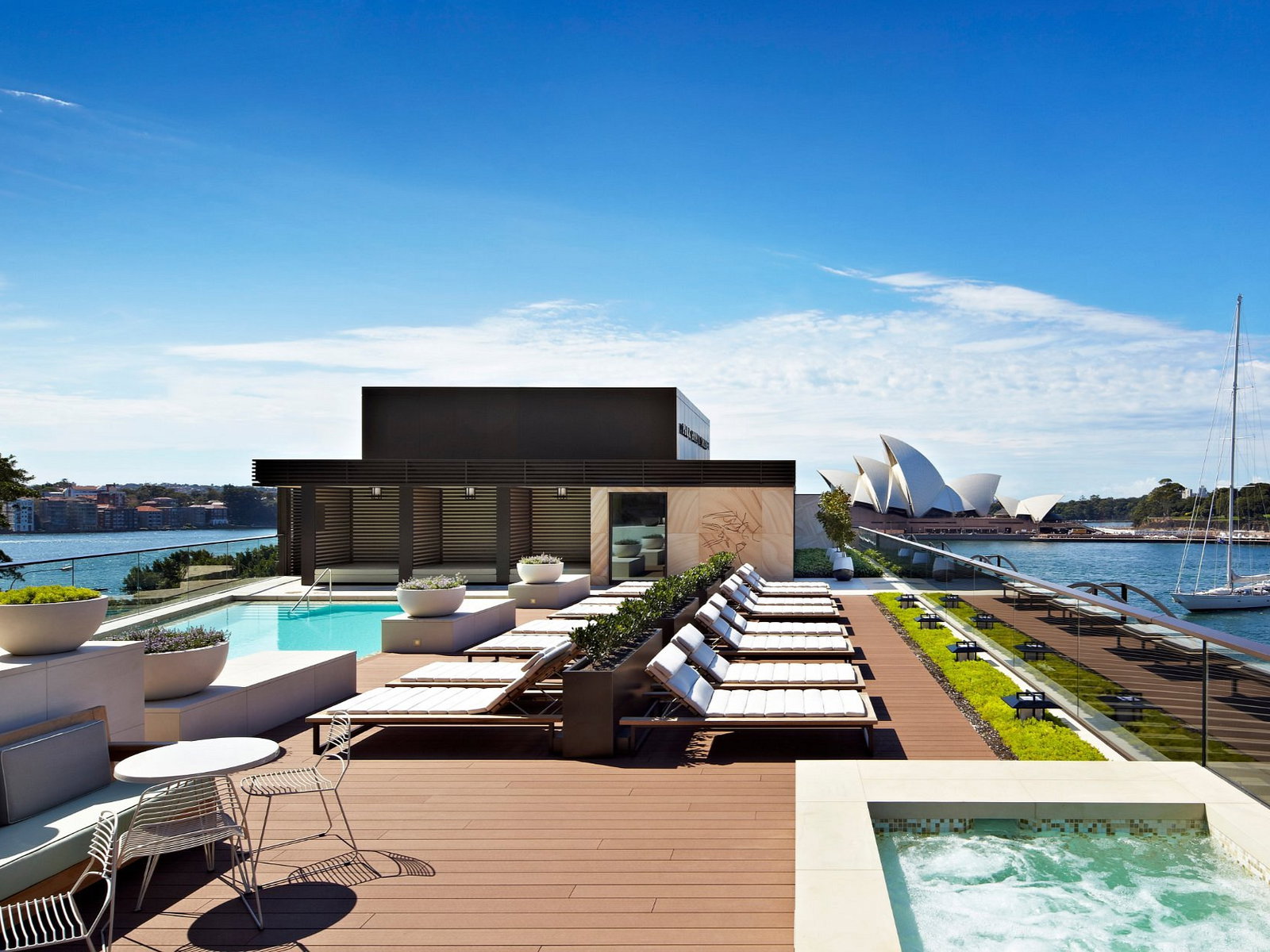 Rooftop pool at Park Hyatt Sydney with lounge chairs overlooks water and a famous architectural landmark. Clear sky, sailboat in the distance.