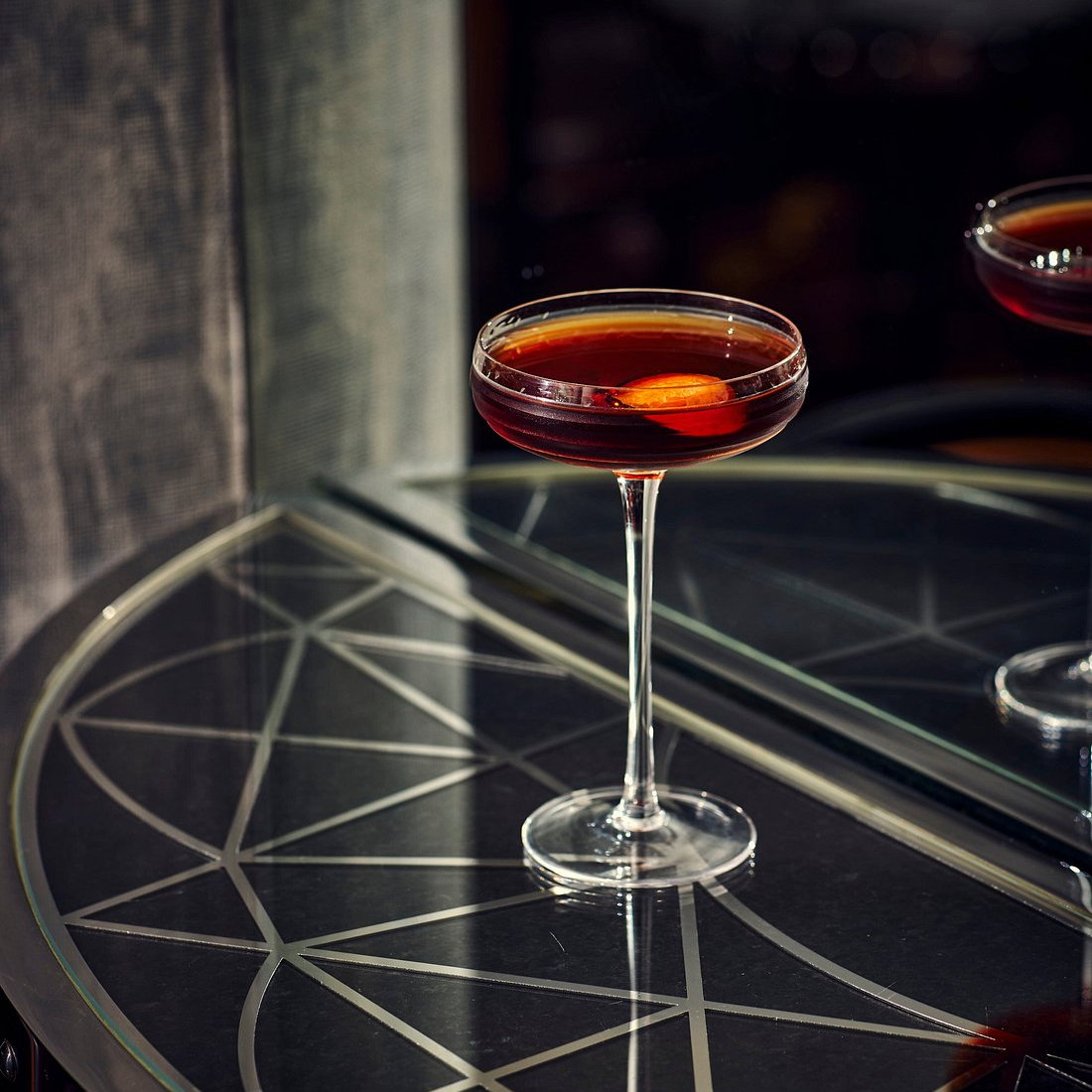 A dark cocktail with an orange slice in a coupe glass sits on a geometric-patterned black table, reflecting a moody, elegant ambiance.