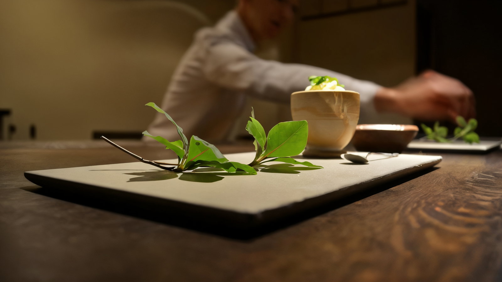 Leaves and a ceramic bowl on a wooden table foreground. Blurred chef in white shirt preparing dish in dim light background. Cozy setting.