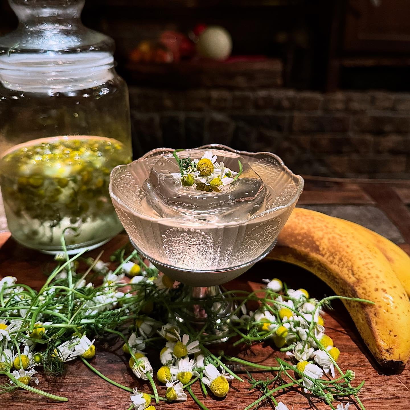 Delicate glass bowl with chamomile float, surrounded by fresh chamomile flowers, a jar of chamomile infusion, and a ripe banana on a wooden table.
