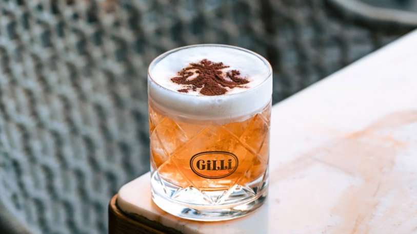 A frothy cocktail in a crystal glass labeled "GiLLi" with cocoa powder design on top sits on a marble table. Woven background pattern.