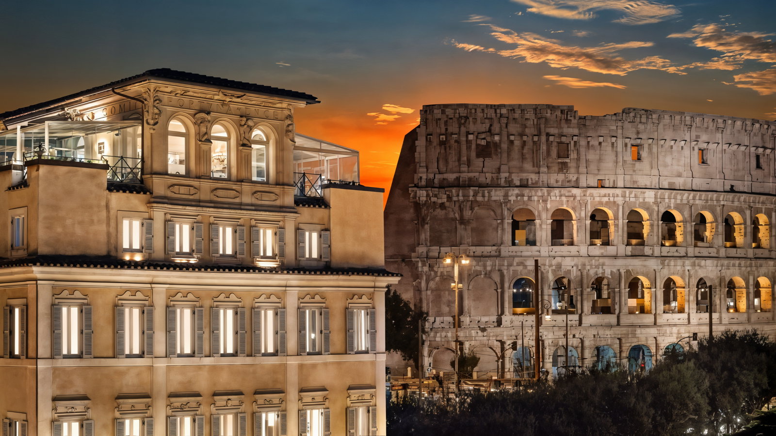 Historic building with lit windows next to the Colosseum at sunset. Vibrant sky with clouds creates a dramatic, warm ambiance.