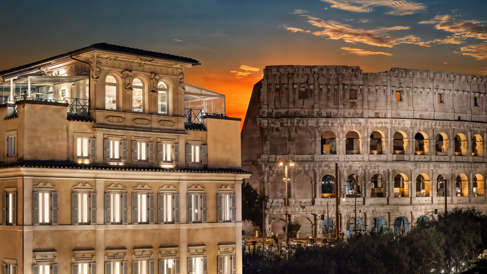 Historic building with lit windows next to the Colosseum at sunset. Vibrant sky with clouds creates a dramatic, warm ambiance.