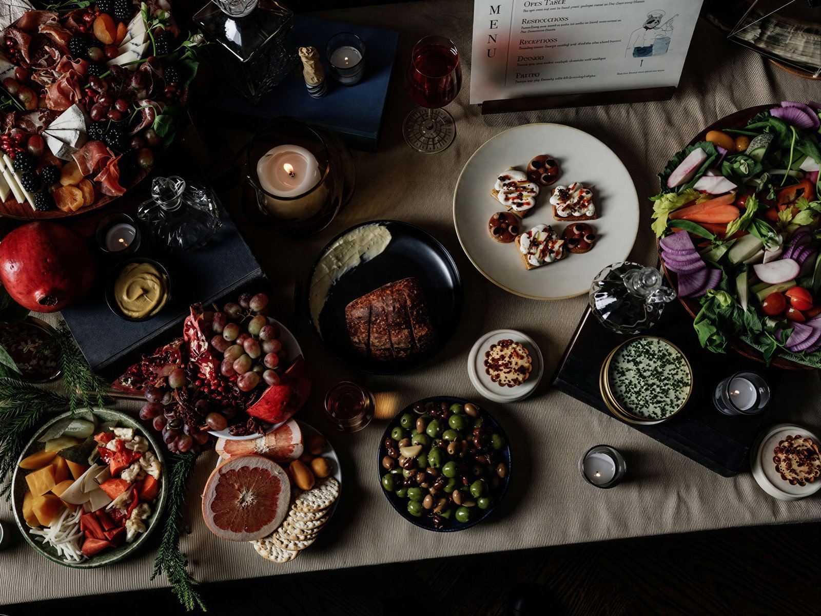 Spread of assorted foods on a dimly lit table with candles. Includes colorful fruits, vegetables, pretzels on cream, and an elegant menu. Cozy mood.