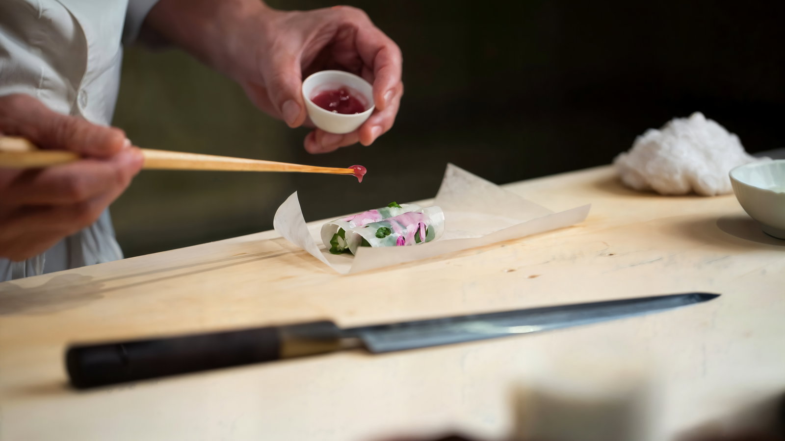 Chef applying sauce to a translucent spring roll on parchment, with a brush and cup. Wooden counter and knife visible.