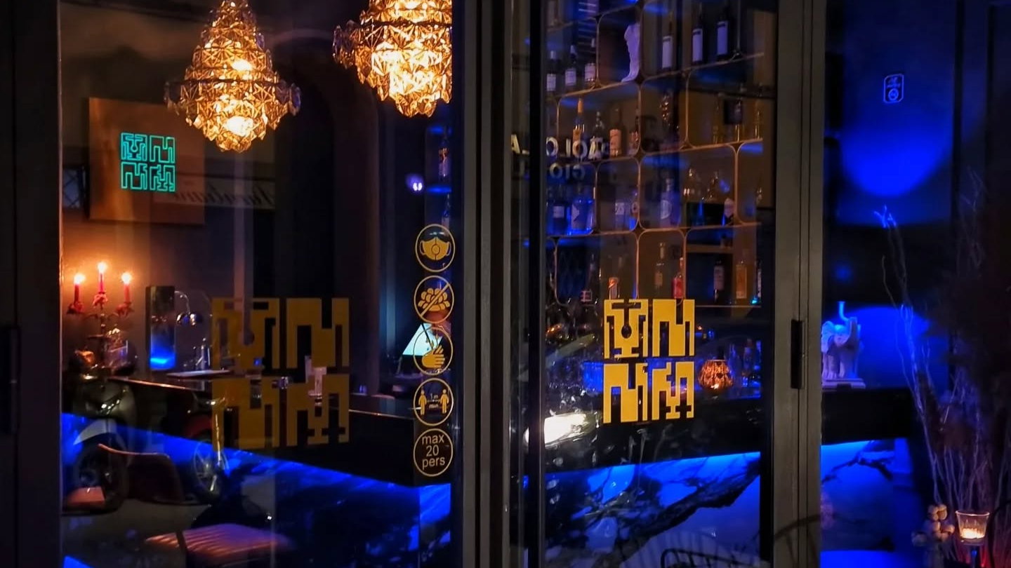 Dimly lit bar with chandeliers, blue glow, and bottles on shelves. Candles on a table near a sign with symbols. Cozy, intimate ambiance.