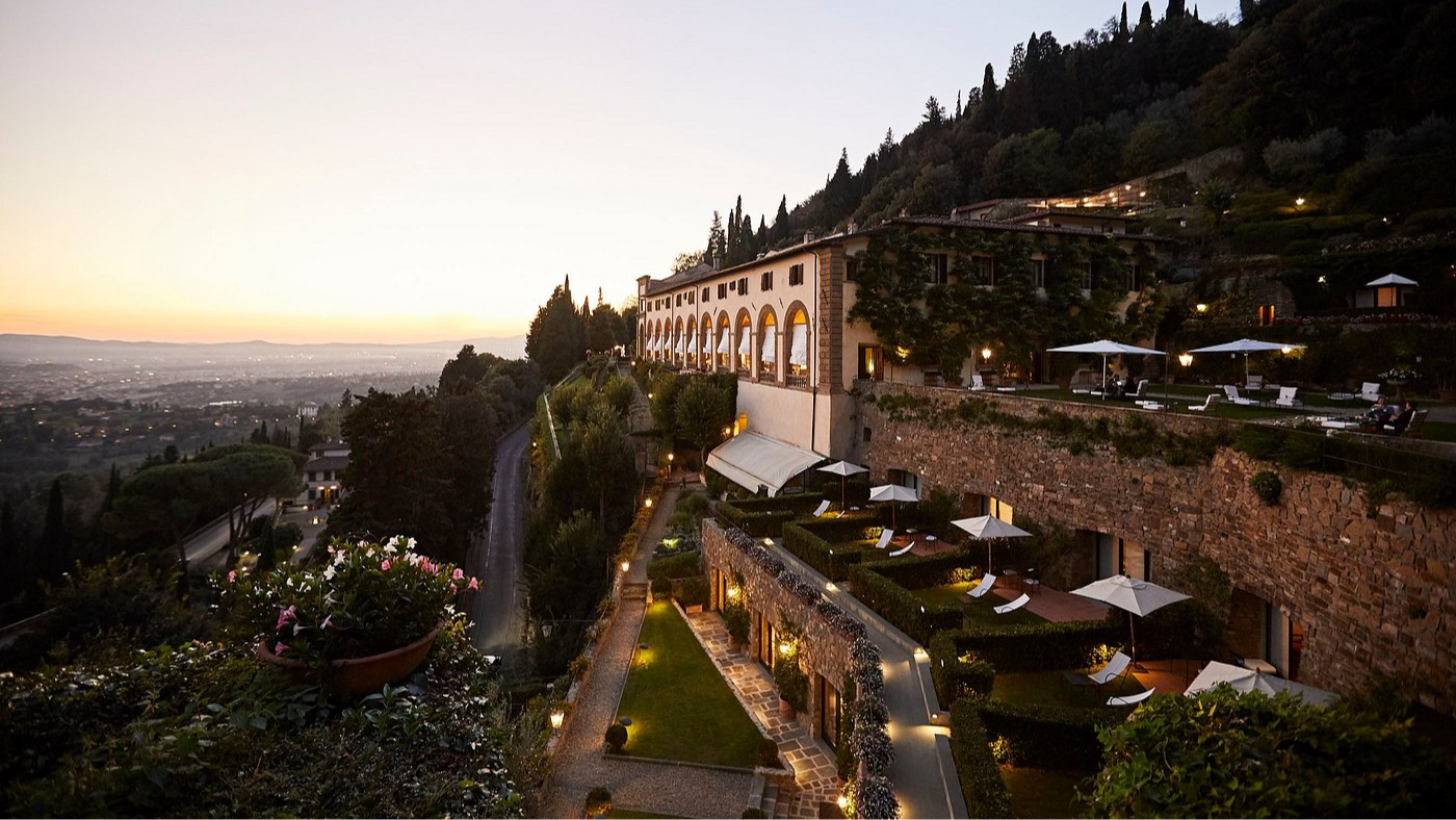 Hilltop hotel at sunset with arched balconies overlooking lush gardens and a distant cityscape, creating a serene atmosphere.