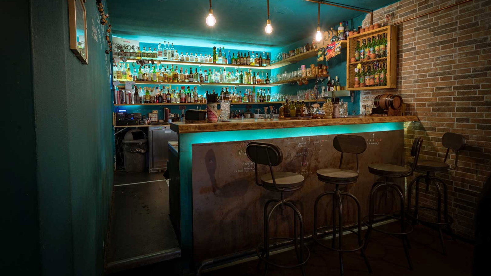 Dimly lit bar with brick walls, teal accents, and three barstools. Shelves filled with bottles and jars glow under ambient light. Cozy vibe.