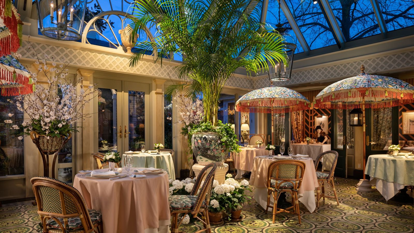 Luxurious restaurant with floral decor, indoor plants, and parasol-like umbrellas. Elegant table settings and a cozy ambiance under a blue sky.