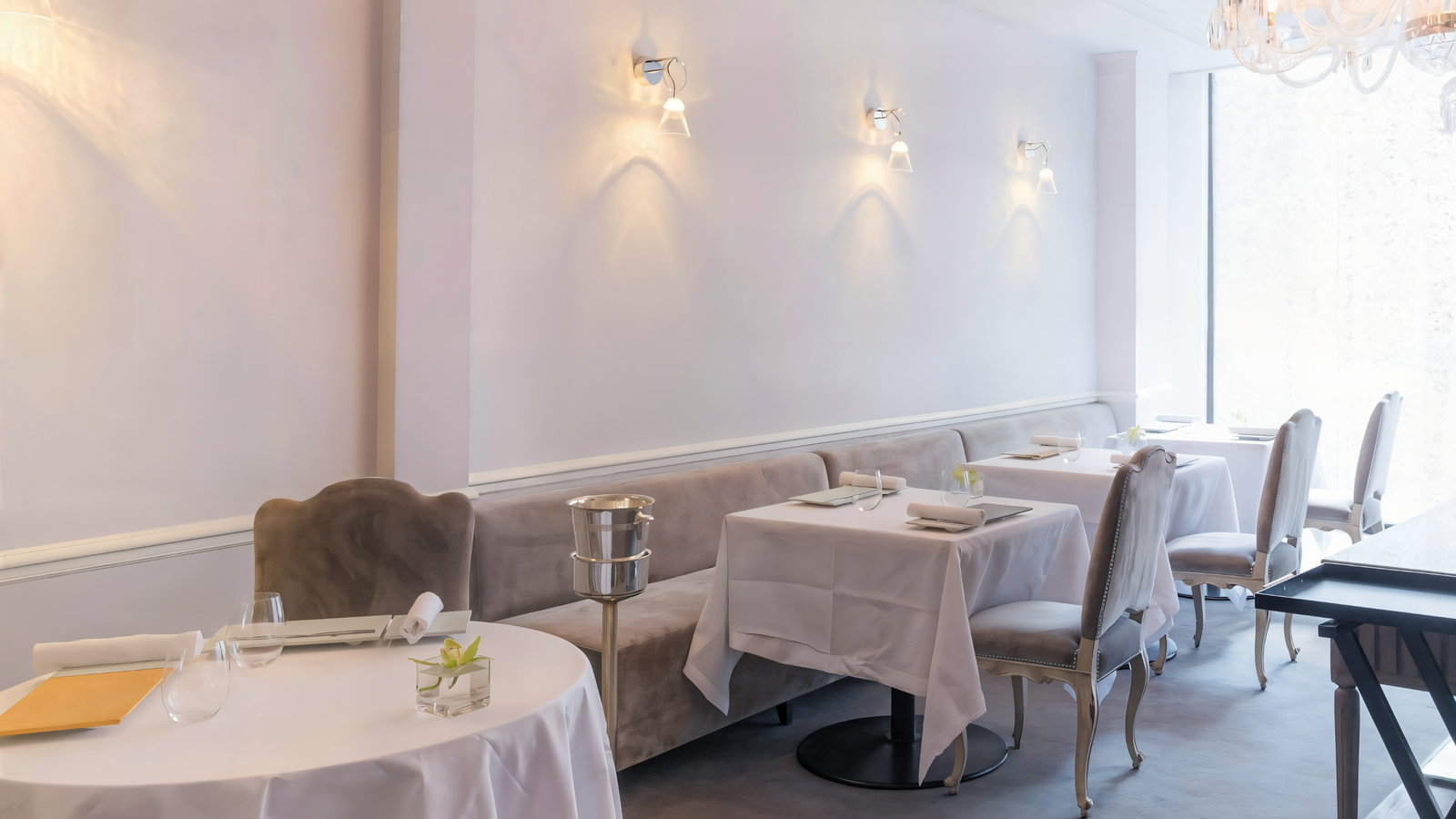 Elegant restaurant with white-clothed tables, plush chairs, and soft lighting. Minimal decor, serene atmosphere, and large window in the background.