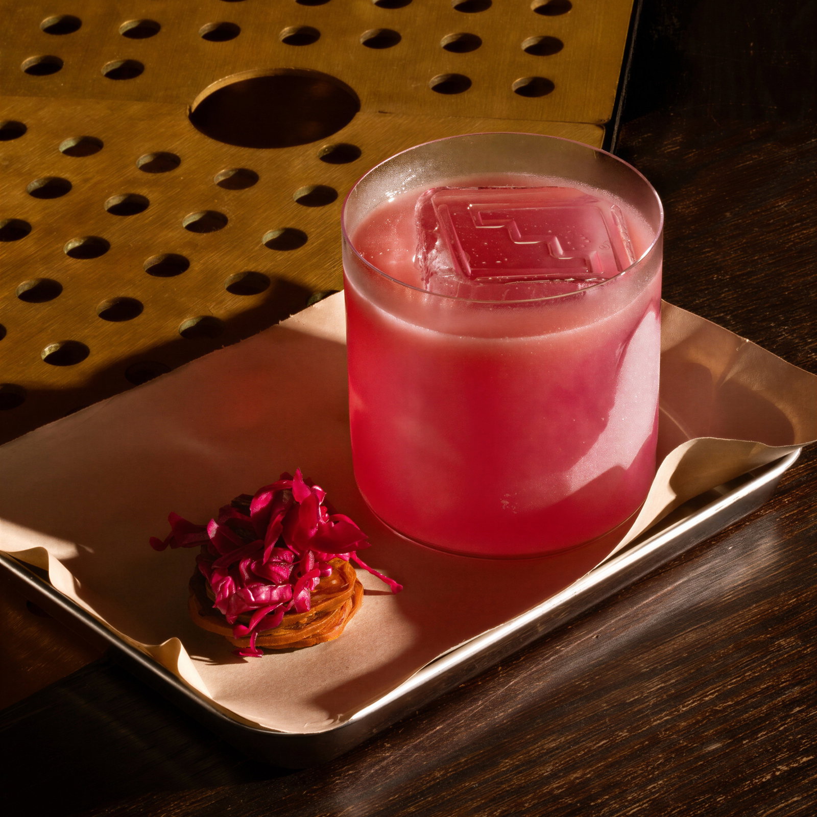 Pink drink in a glass with a large ice cube on a tray. Garnished with red flowers. Background has a brass textured surface.