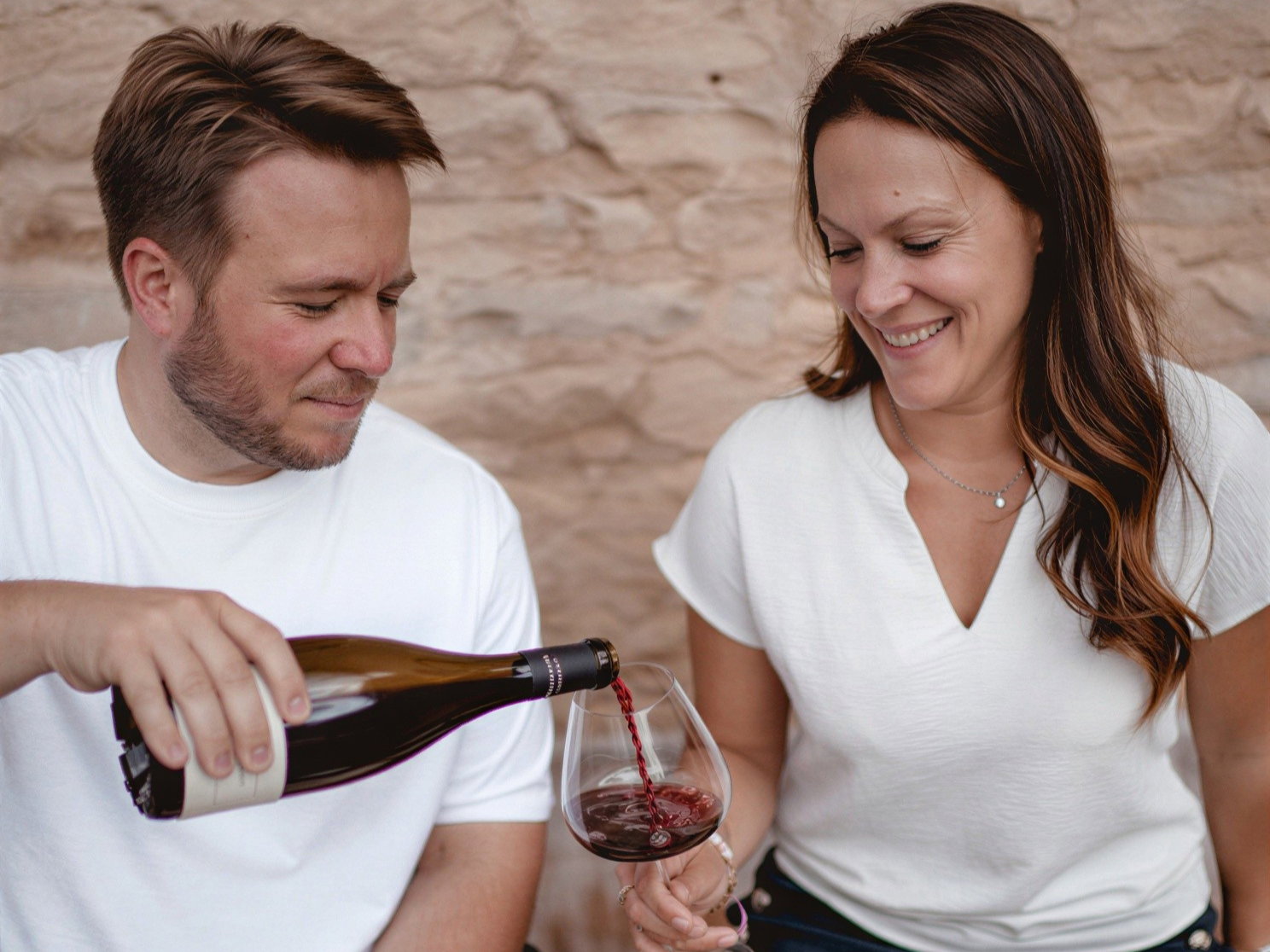 Antoine Amiot-Servelle in white shirt pours red wine for Prune Amiot-Servelle in shorts and white top. They are seated against a stone wall, creating a relaxed mood.