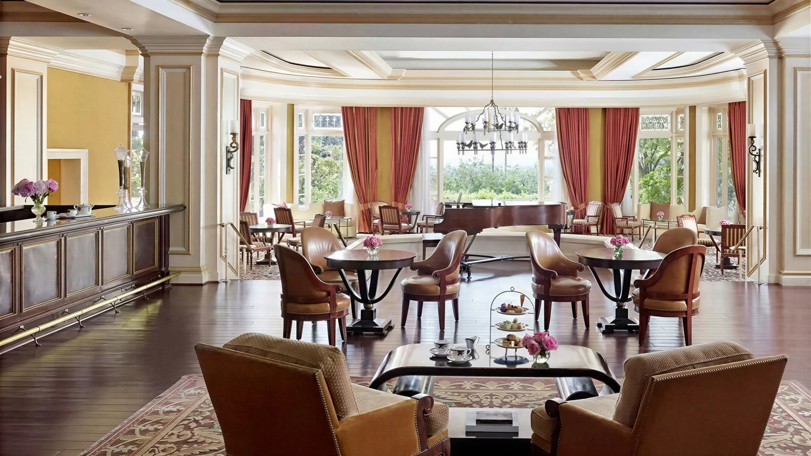Elegant tea room with plush chairs, tables, and a grand piano. Large windows with red curtains overlook a garden, creating a cozy ambiance.