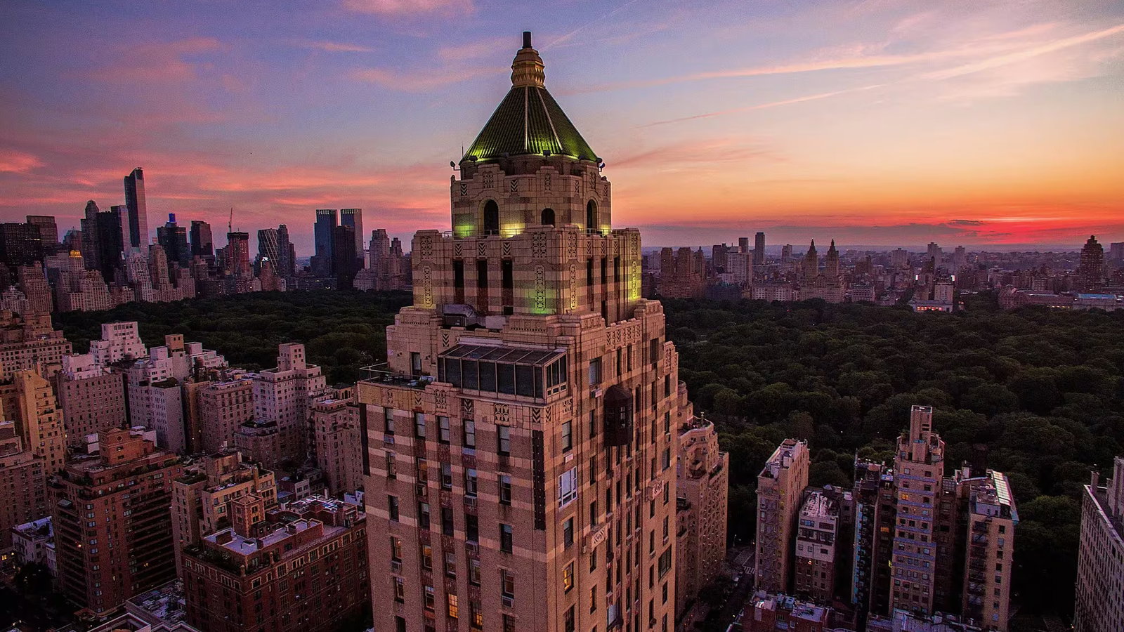 A tall, ornate building with a green-lit top stands against a vibrant sunset. Central Park's greenery and a distant city skyline complete the scene.