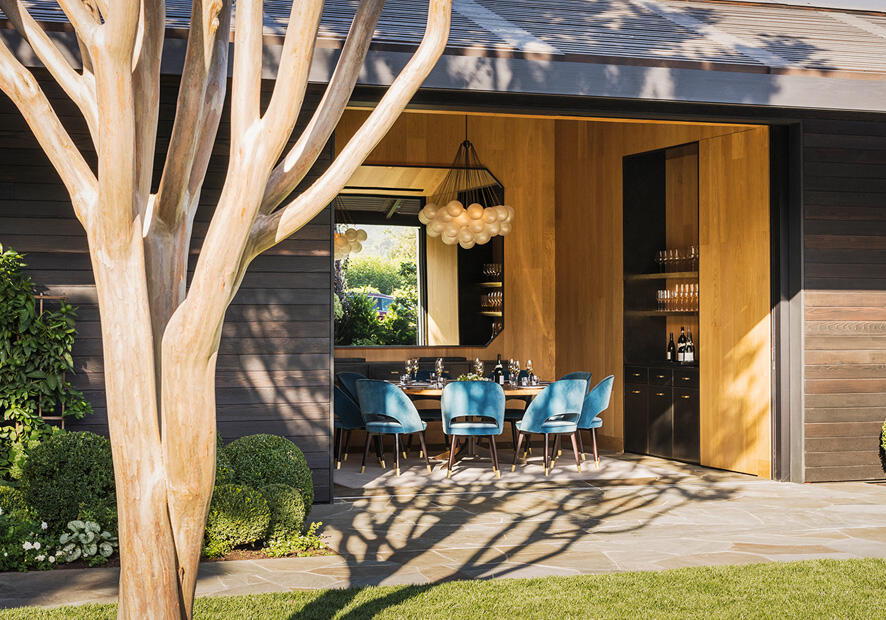 A tree is in the forefront of an open concept private dining room with blue chairs and a bottle of wine.