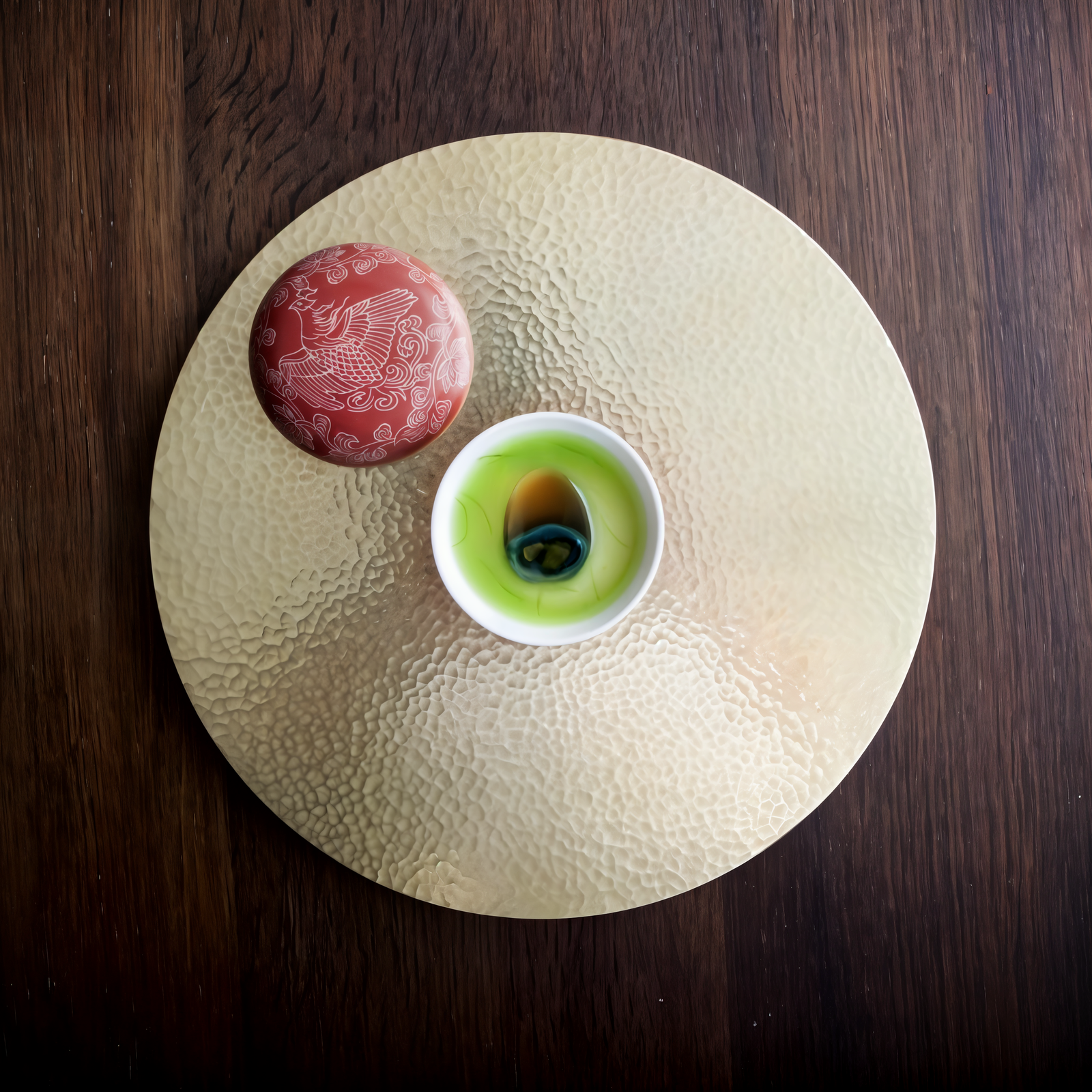 Ceramic cup with green tea sits on a textured golden plate. A red lid with white patterns lies nearby. Wood surface backdrop.