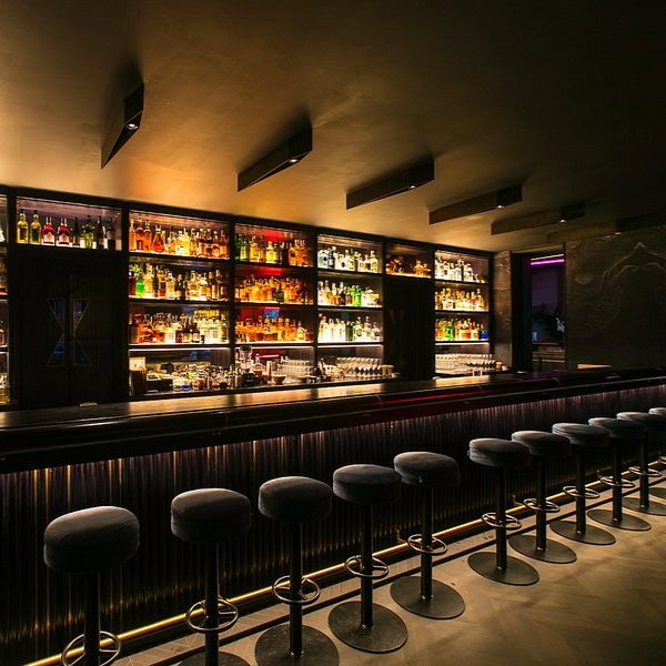 Dimly lit bar with a long counter, barstools, and shelves full of colorful liquor bottles. Warm ambiance with sleek, modern design.