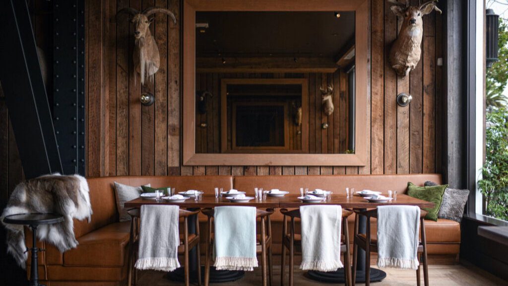 Rustic dining room with wooden walls, large mirror, and taxidermy mounts. Table set for eight with white cloths and leather seating.