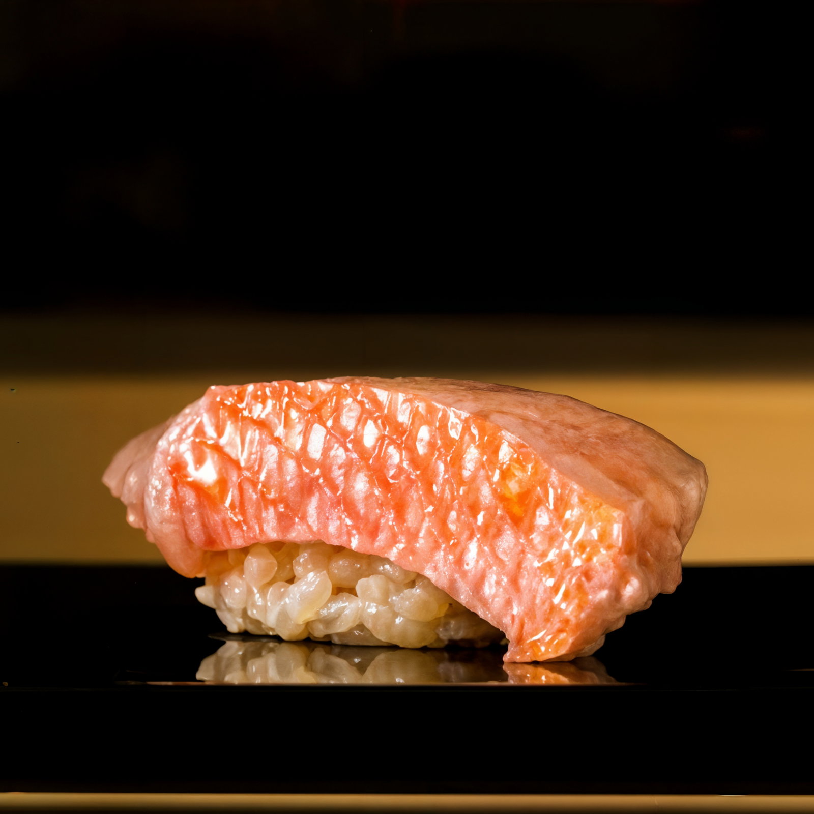 Close-up of a piece of sushi with glossy pink fish atop rice on a shiny black surface, set against a dark blurred background.