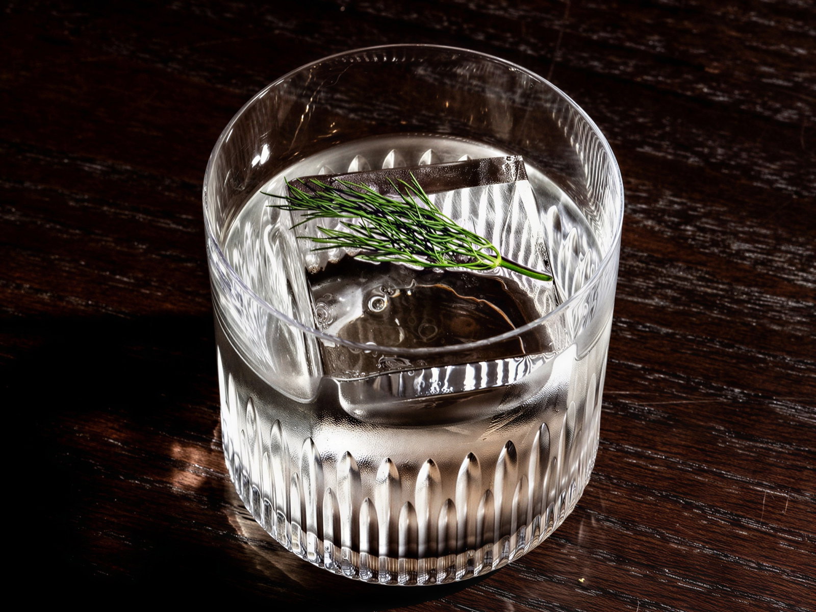 A clear glass with a large ice cube and dill sprig on a dark wooden surface. The setting is moody with visible reflections on the table.