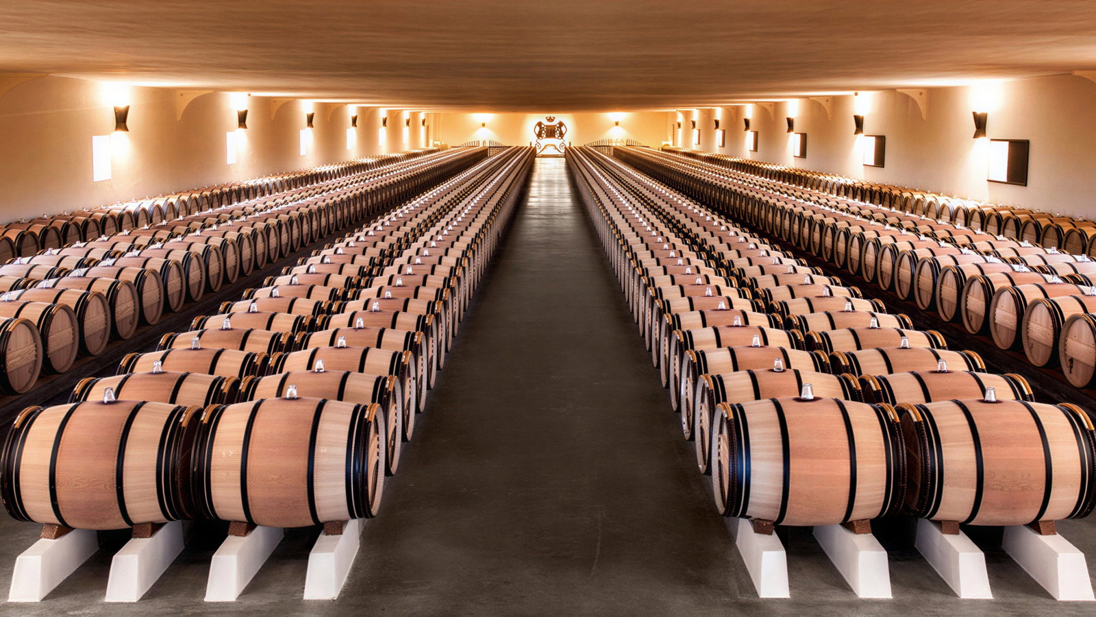 Rows of wooden barrels line a dimly lit wine cellar, creating a symmetrical pattern. Warm light illuminates beige walls, evoking a serene mood.