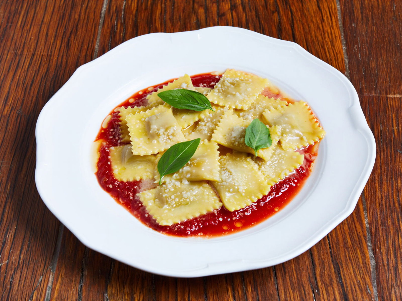 My Grandmother's Ravioli from Locanda Verde with tomato sauce and basil on a white plate, sprinkled with cheese, placed on a wooden table. Warm, inviting setting.