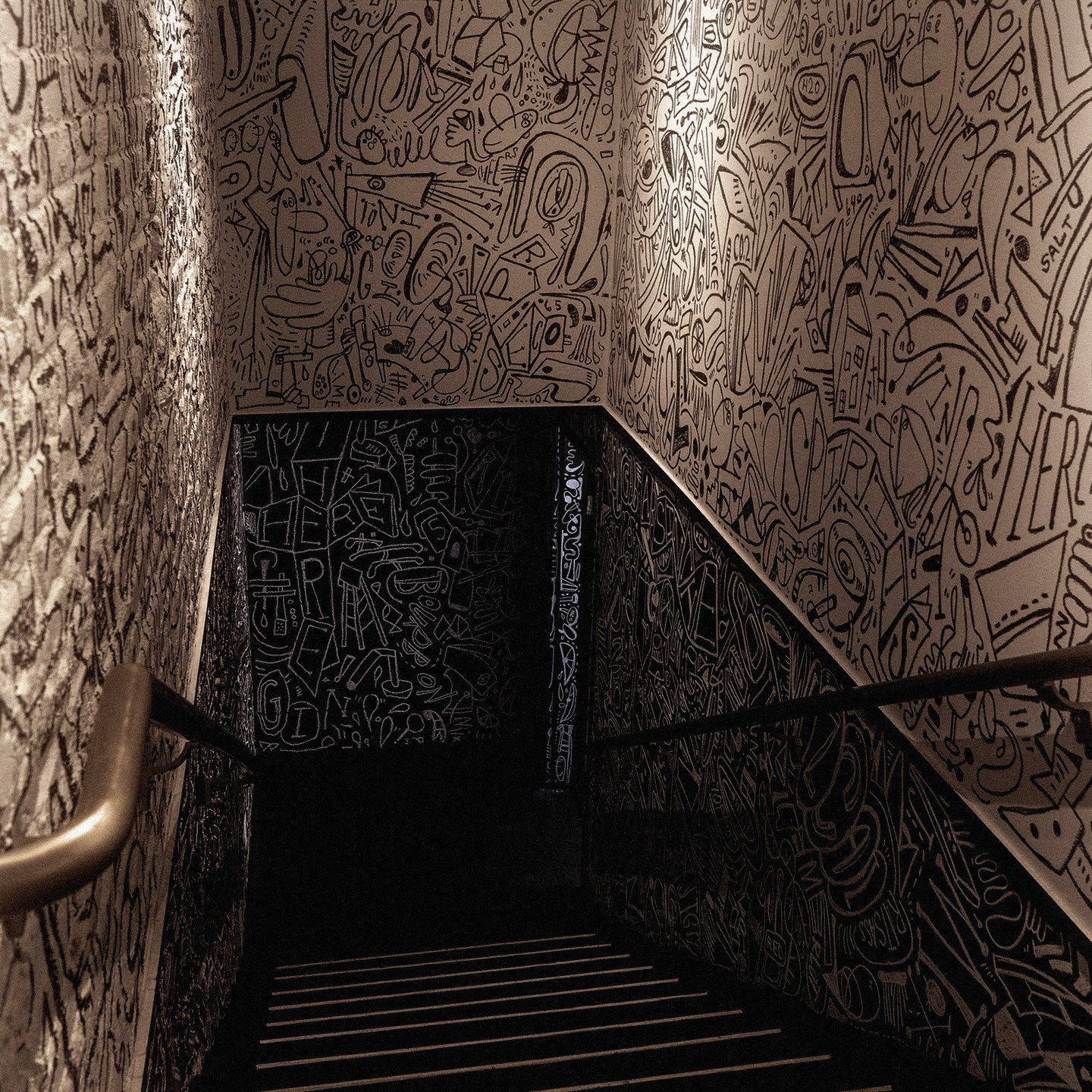 Staircase with intricate black and white graffiti-style doodles on walls. A handrail on the left adds depth. Dimly lit, urban vibe.