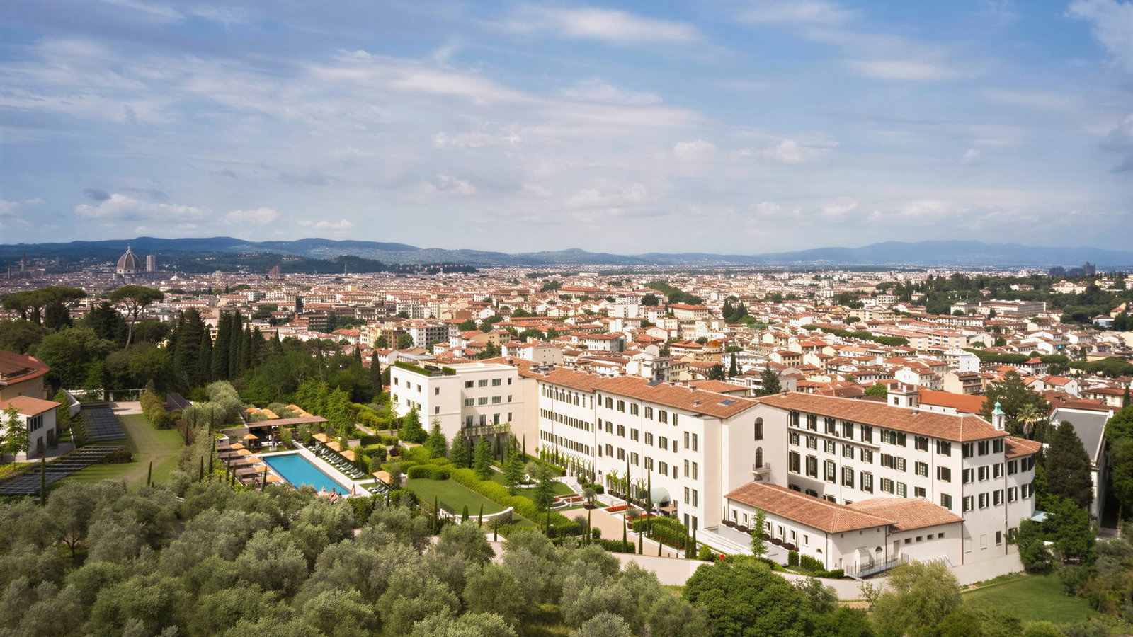 Aerial view of Collegio alla Querce and Florence with white buildings, a pool, and lush green gardens in the foreground. Red-roofed cityscape and distant mountains.