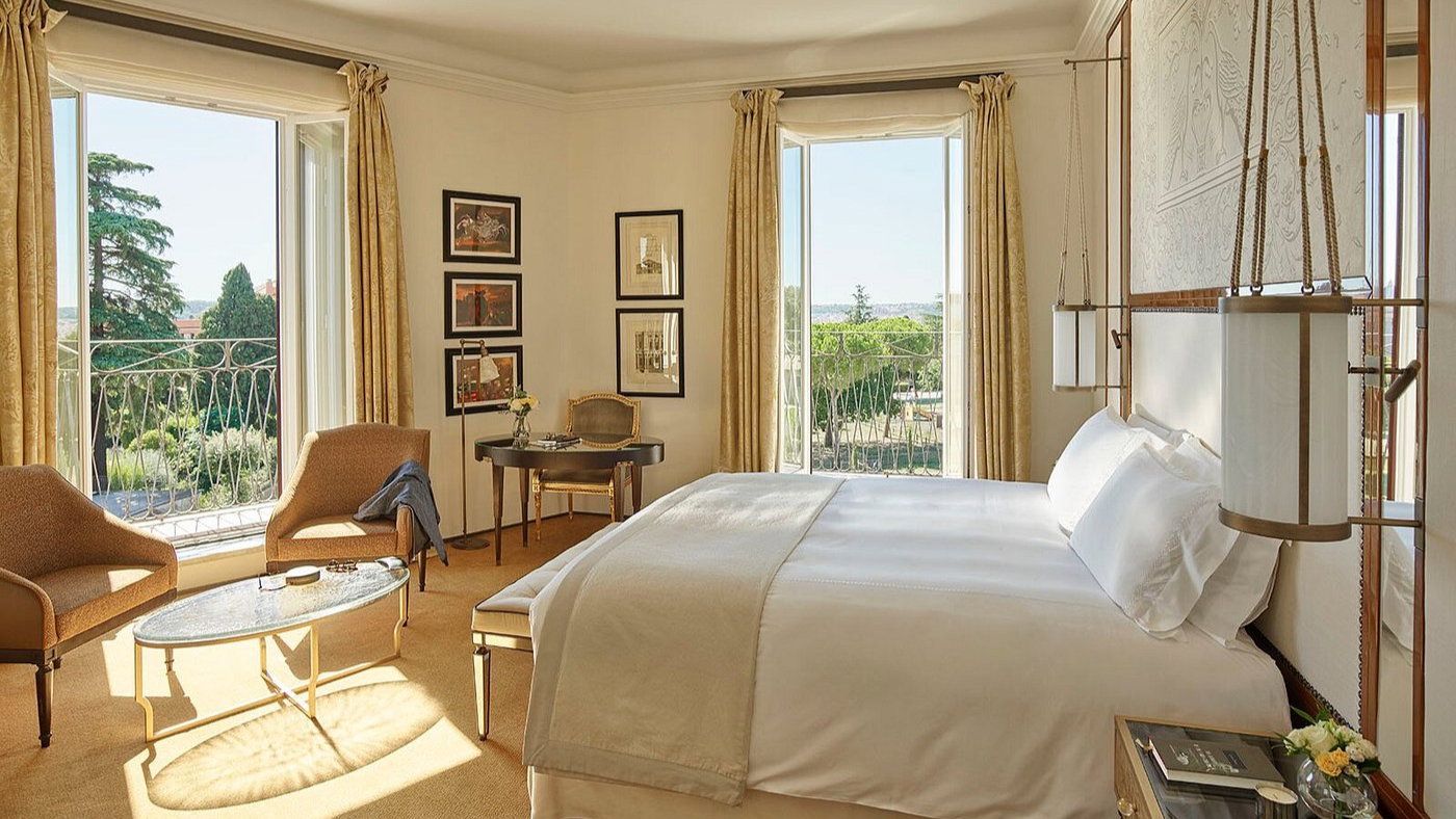 Elegant hotel room with a large bed, two armchairs, and a glass table. Sunlit through open windows with garden views. Calm and luxurious.