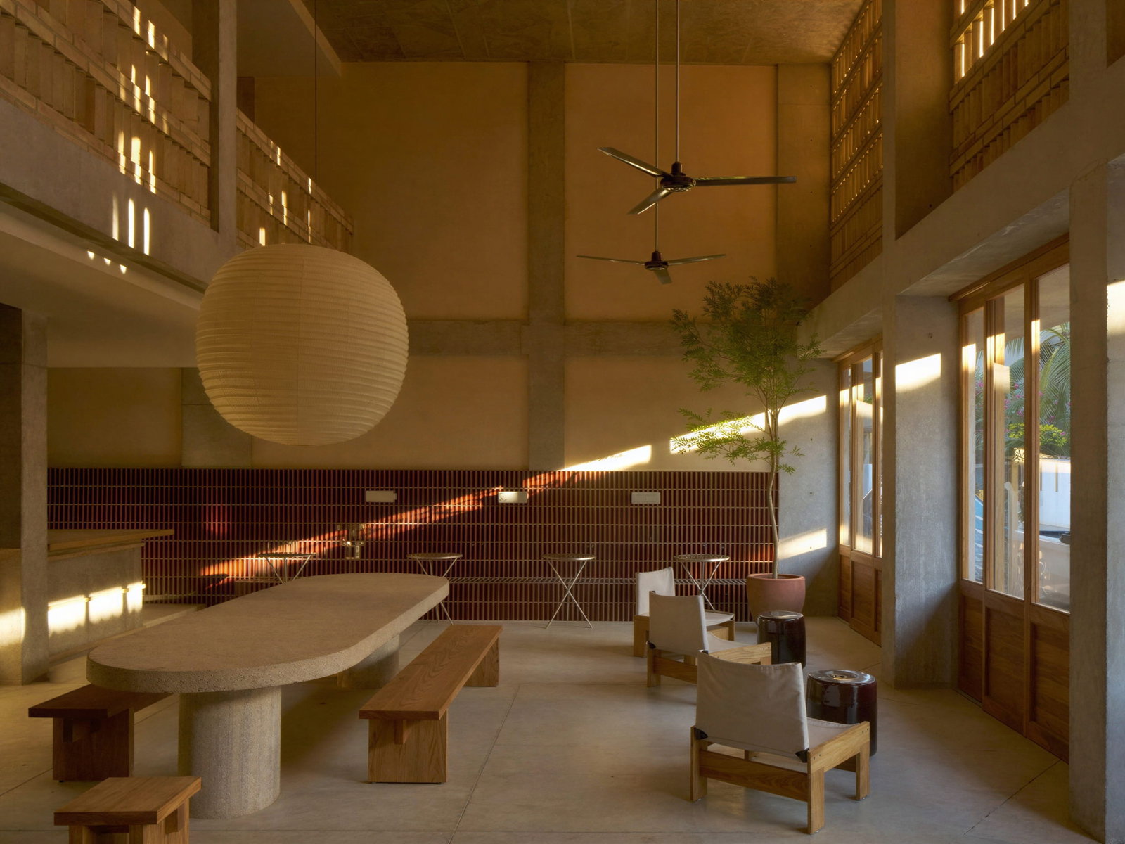 Spacious coffee bar room at Hotel Humano with large table, benches, paper lantern, and ceiling fans. Sunlight streams through windows, casting warm patterns. Tranquil mood.
