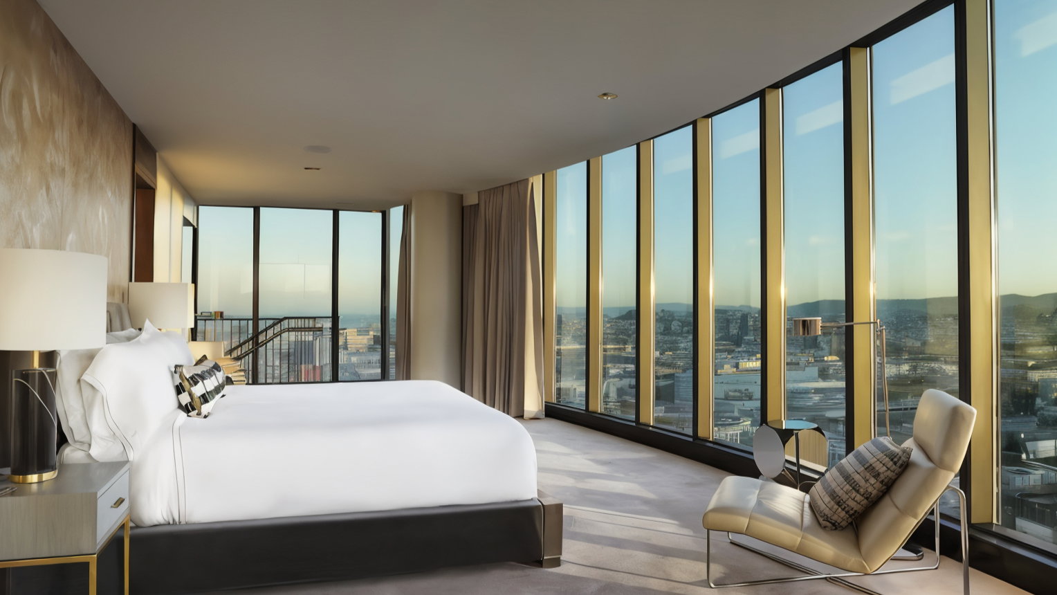 Luxurious bedroom with a white bed, modern chair, and city view through large windows. Warm tones and elegant decor create a serene mood.
