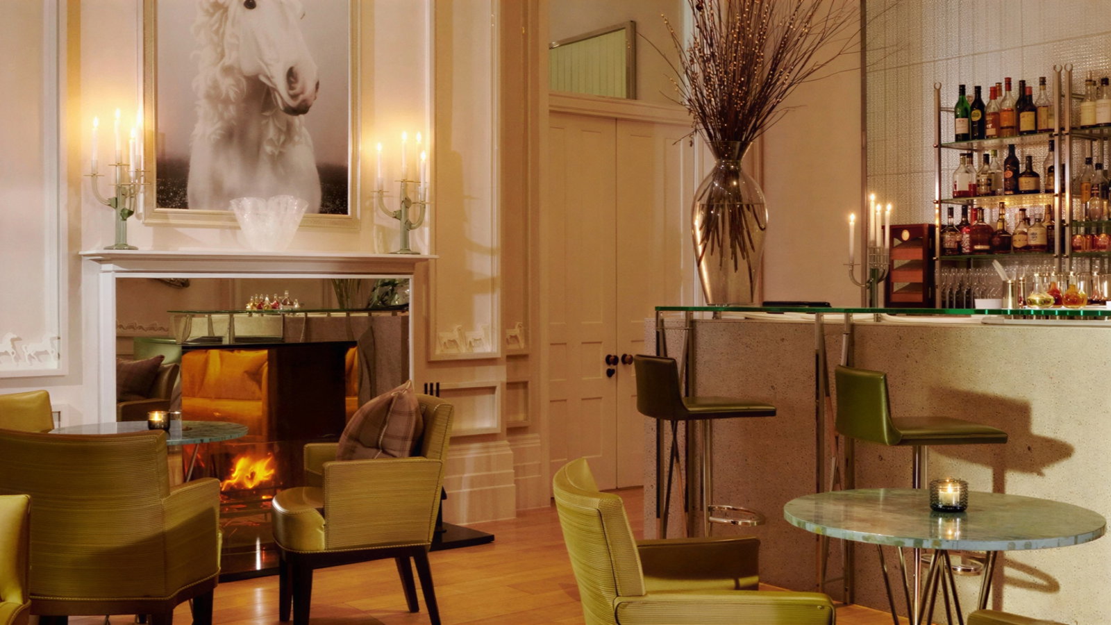 Elegant lounge with green chairs, a lit fireplace, and a bar. A white horse portrait and candles enhance the cozy atmosphere.