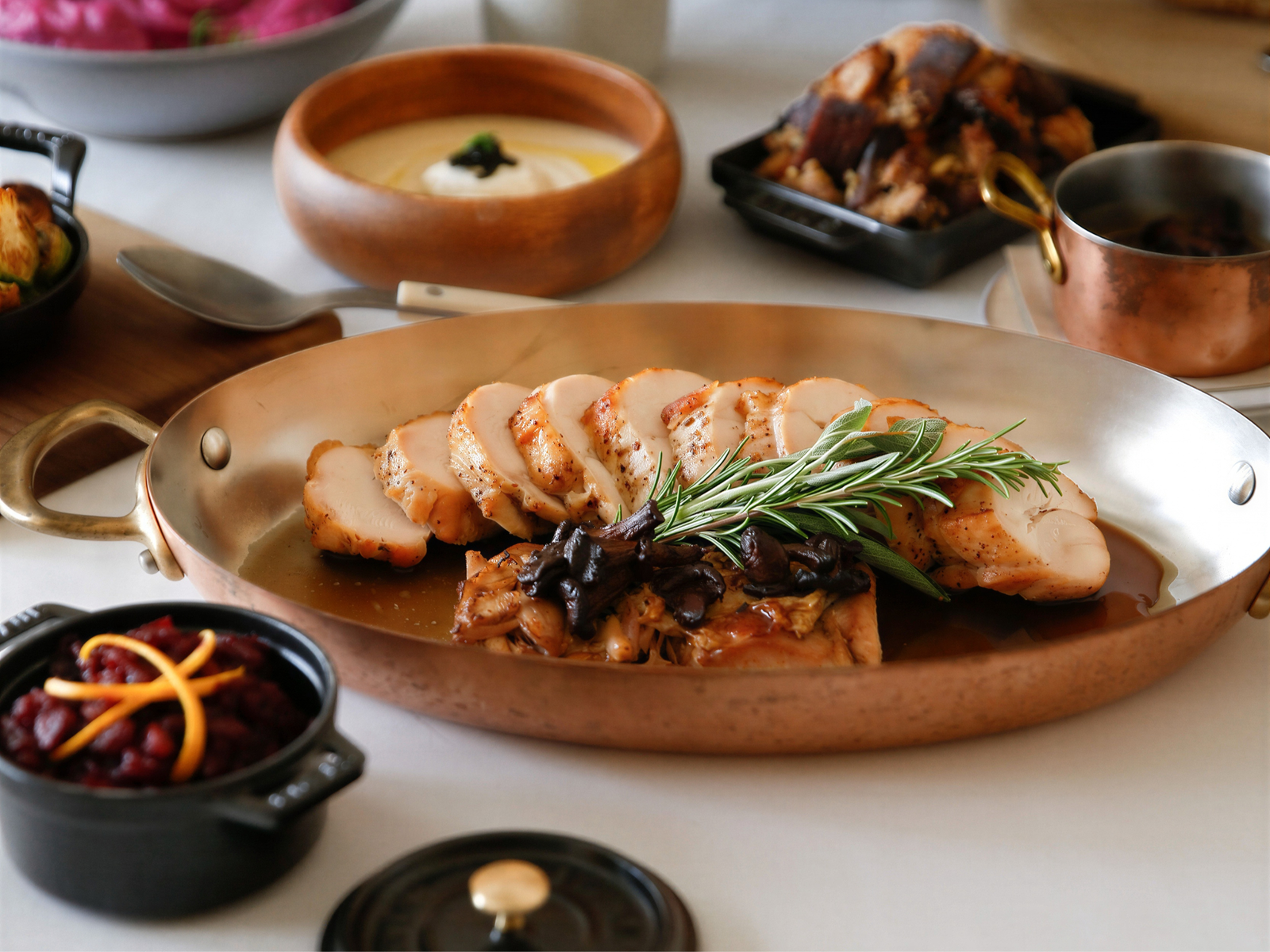 Sliced turkey breast with rosemary and sauce on a platter, surrounded by bowls of cranberry sauce, gravy, and side dishes on a table.