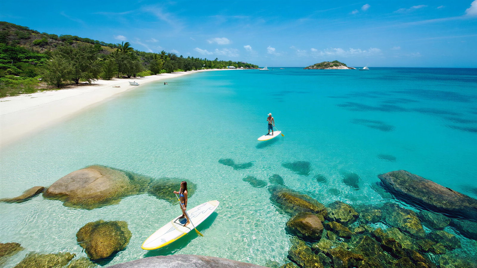 Paddleboarding at Lizard Island Resort at Lizard Island Resort on clear turquoise water near a sandy beach with lush greenery. Rocks are visible underwater, and a boat is in the distance.