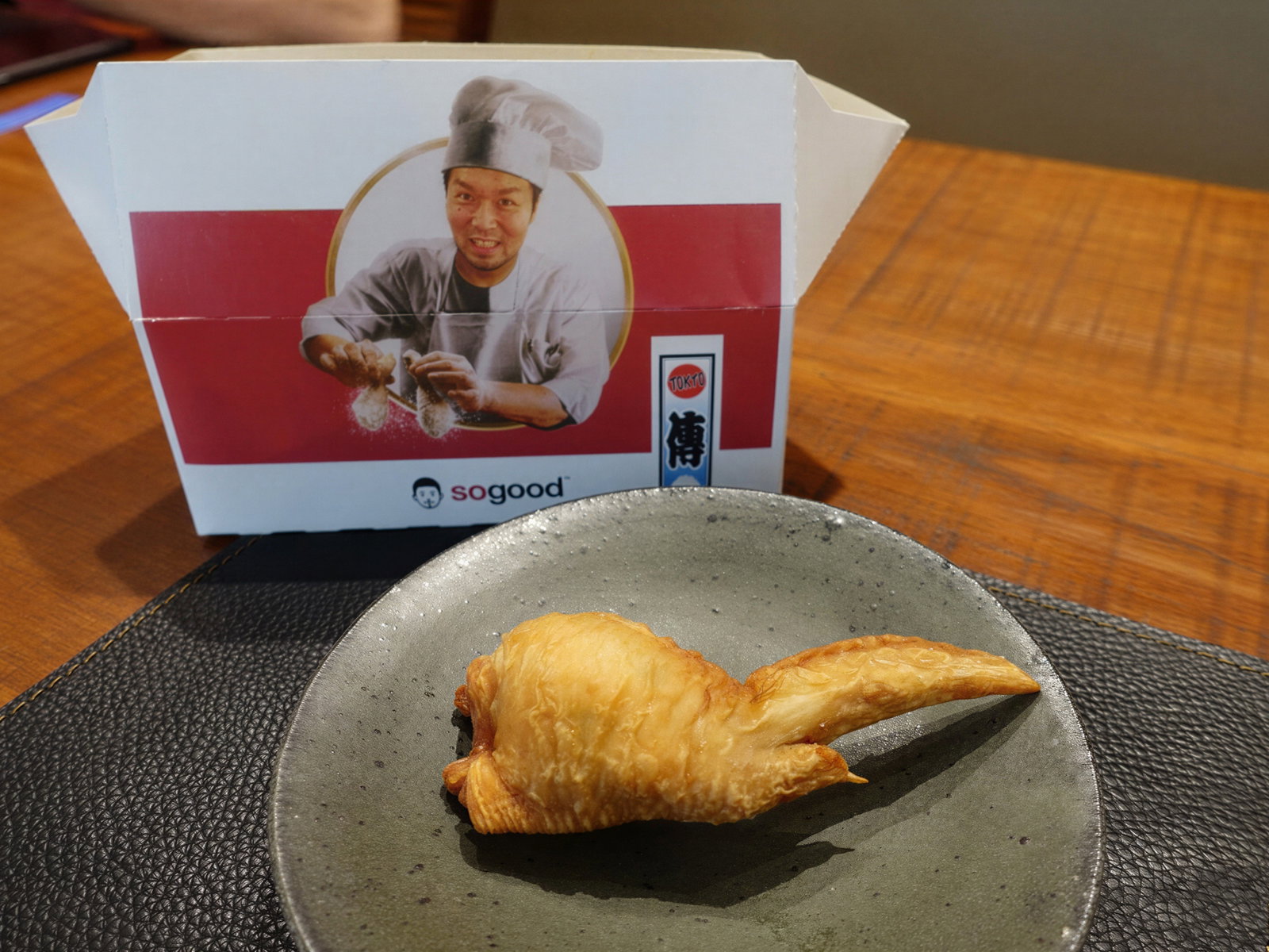 A plate with a golden pastry resembling a fish is on a table. Behind it is a box featuring a chef, flouring hands, with "sogood" text.