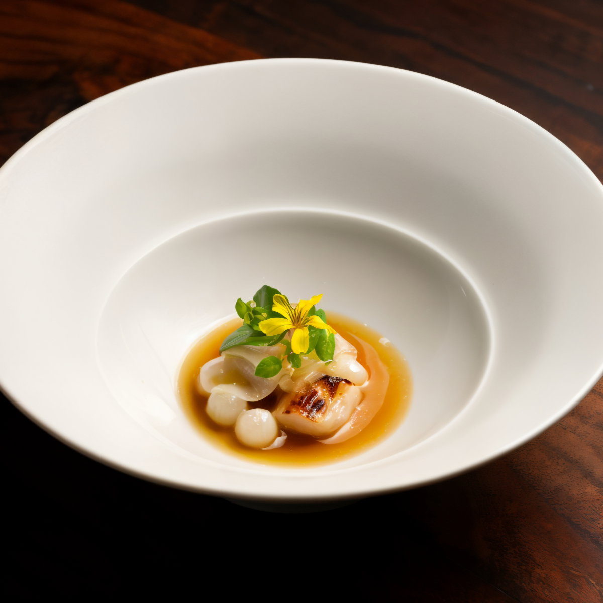 Gourmet dish with scallops in broth, topped with greens and a yellow flower, served in a white bowl on a dark wood table.