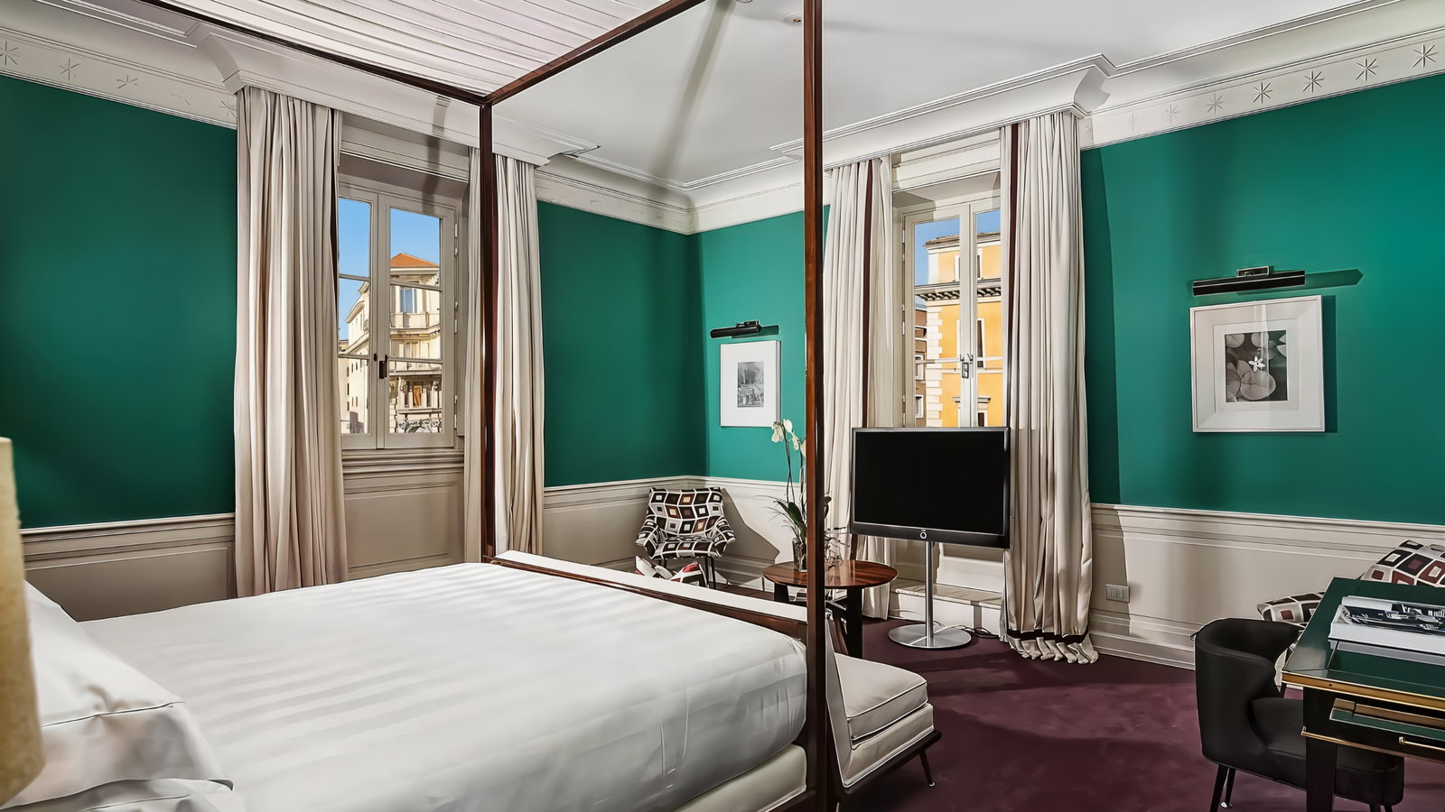 Elegant bedroom with a four-poster bed, green walls, and a large window revealing a sunny building. Modern furniture and framed art enhance the decor.