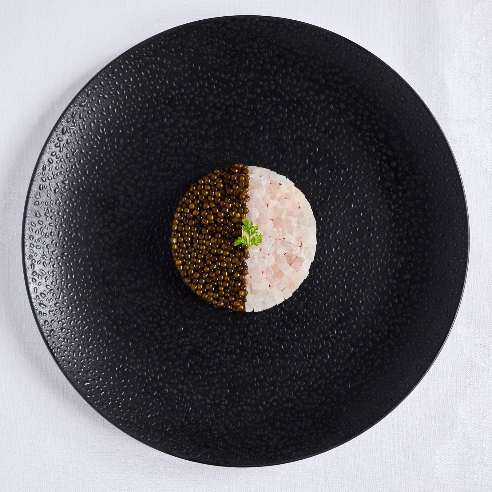 Round dish features half caviar, half diced fish, garnished with a sprig on a textured black plate. Elegant, minimalist presentation.