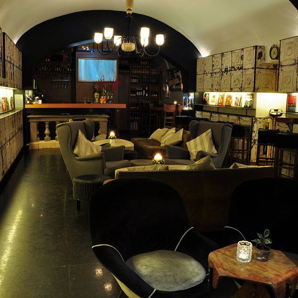 Cozy bar with armchairs and soft lighting, vintage patterned walls, books on shelves, and a dimly lit countertop, creating a warm ambiance.