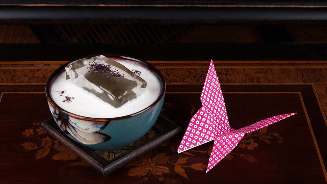 A ceramic bowl with white foam and translucent ice, topped with purple flakes, sits on a wooden table beside a pink origami crane.