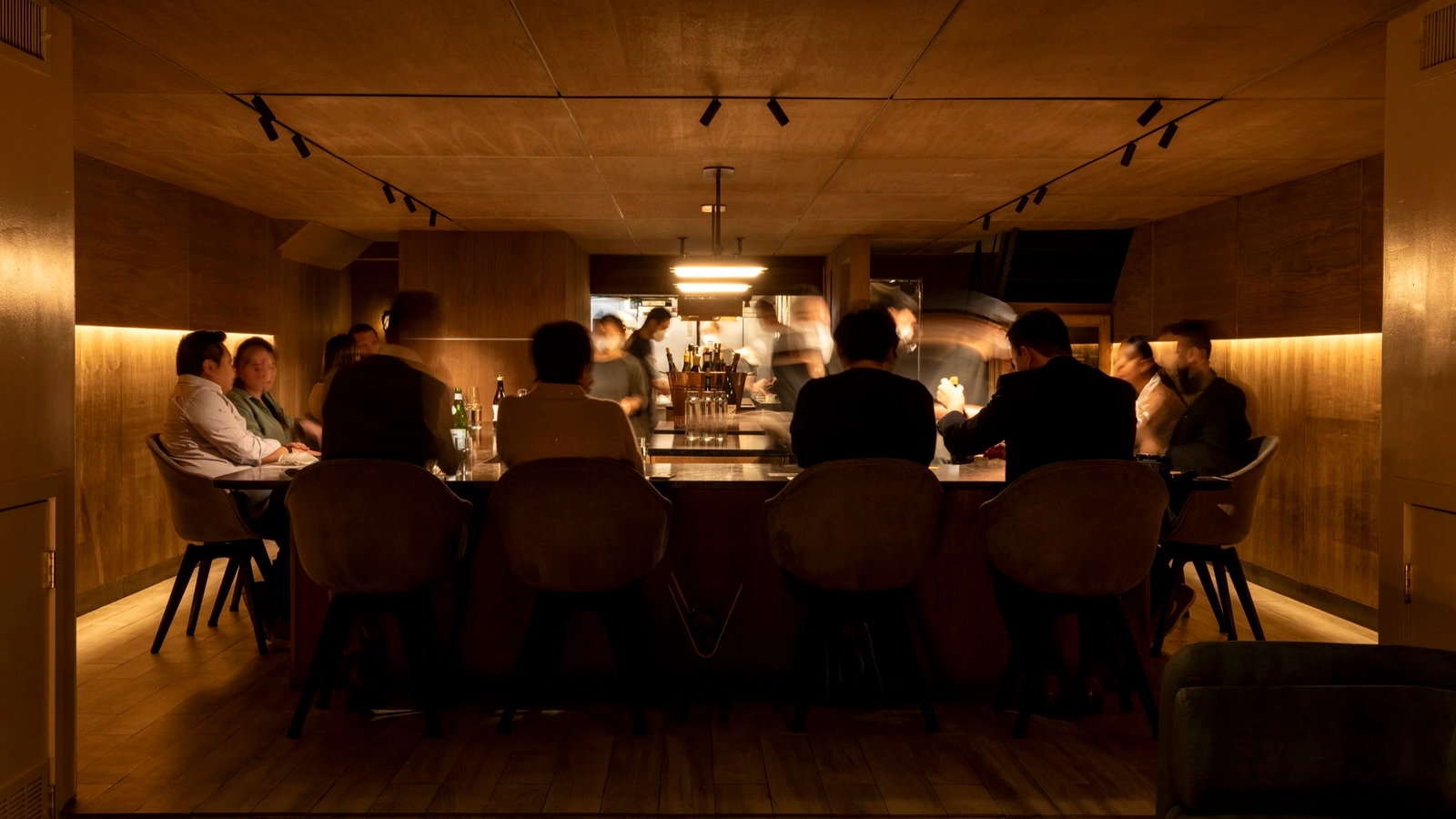 Dimly lit bar with people seated around a counter enjoying drinks. Bartenders in motion behind them. Warm wooden interior creates a cozy mood.