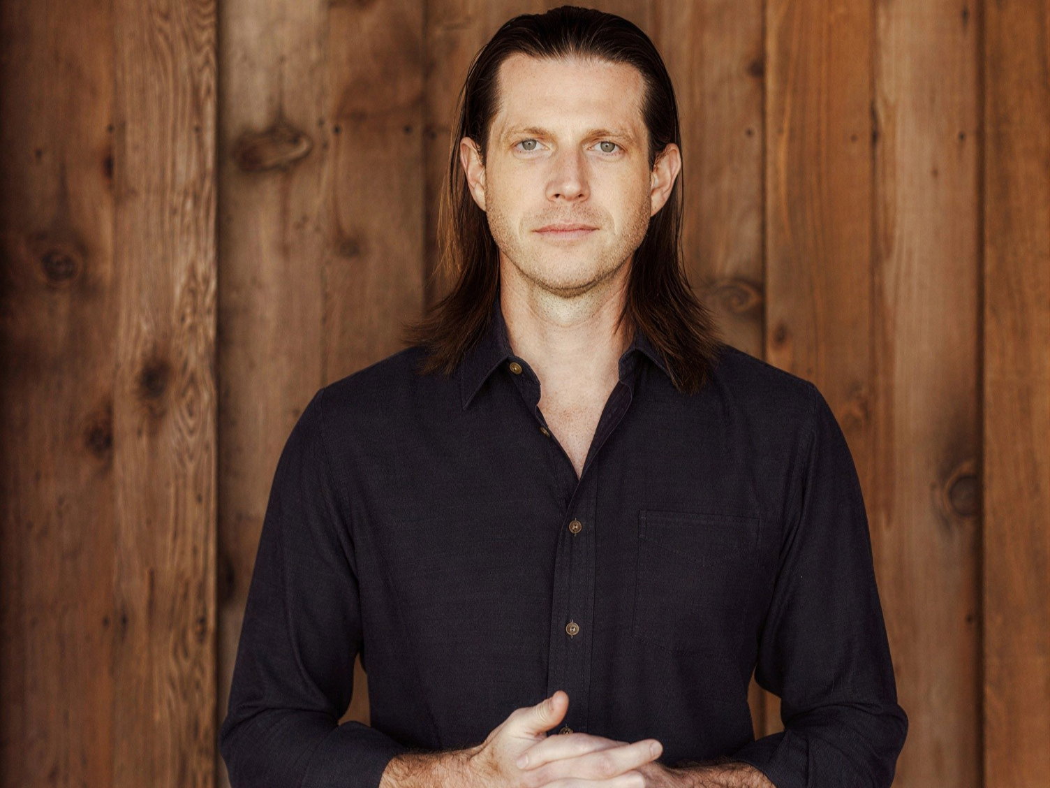 Will Harlan with long hair and a neutral expression stands in front of a wooden wall, wearing a dark shirt. The mood is calm and composed.
