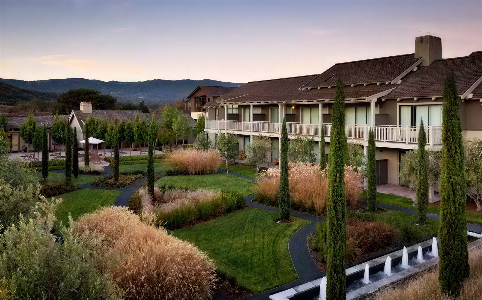 Green garden with tall trees, a winding path, and a fountain in front of a two-story building. Mountains and calm sky in the background.