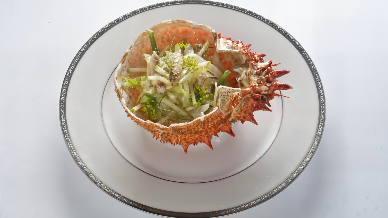 Crab shell filled with shredded vegetables and herbs on a silver-edged plate. The setting is minimalist with a white background.