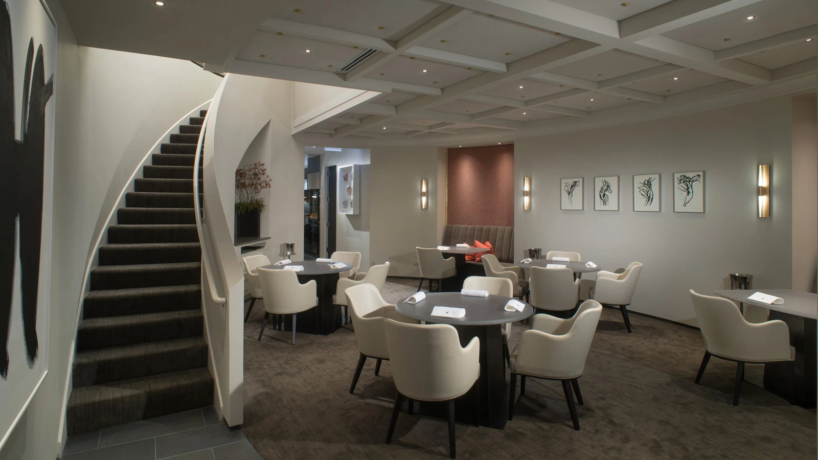 Elegant restaurant interior with round tables, cream chairs, and art on walls. Curved staircase on left, dim lighting creates cozy ambiance.