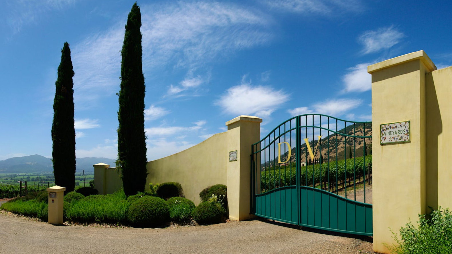 Tall green gates with "DV" stand before a vineyard. Two tall cypress trees and lush greenery surround. Blue sky and mountains in the background.