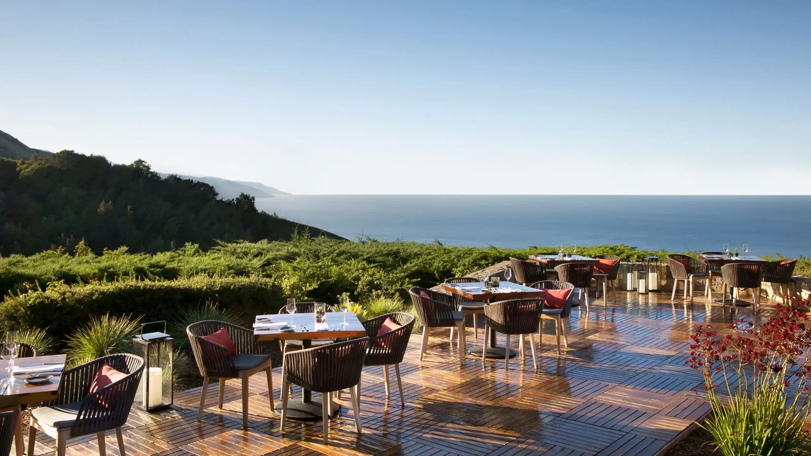 Outdoor patio with empty dining tables and chairs overlooks ocean and hills. Wooden flooring, lush greenery, clear sky, and tranquil vibe.