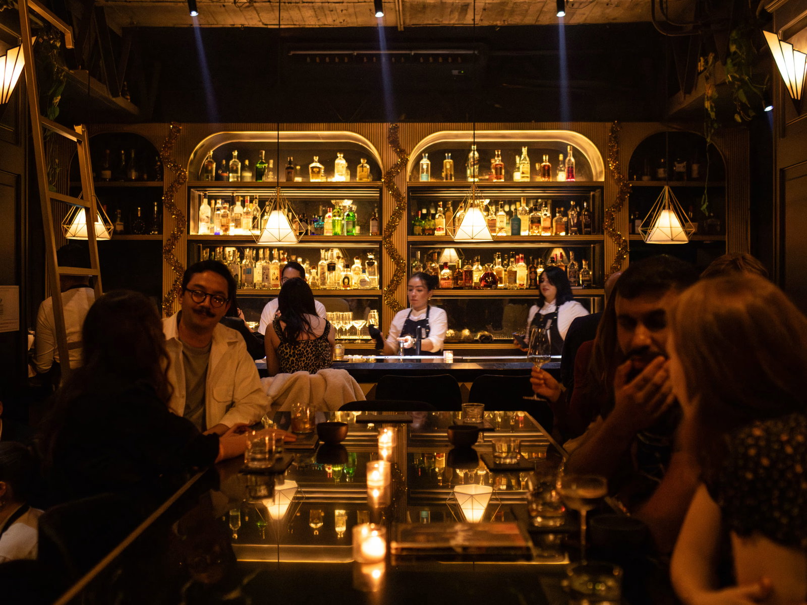 People sit at a dimly lit bar with glowing candles and shelves of bottles behind. Bartenders serve drinks, creating a warm, lively atmosphere.