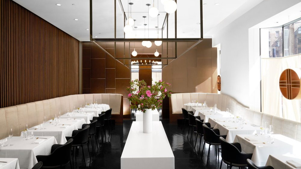 Elegant restaurant interior with white tablecloths, black chairs, wood paneling, and floral centerpiece. Bright lighting and modern decor.