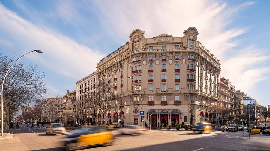 Historic beige building with red accents on a busy street corner; blurred taxis pass by. Blue sky, bare trees line the sidewalk.